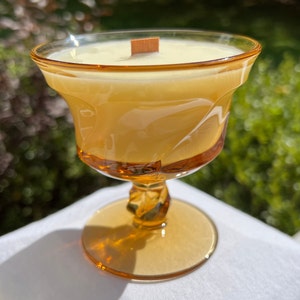 May include: A clear glass candle holder with a amber tint and a wooden wick. The candle holder is shaped like a goblet and has a wide base.
