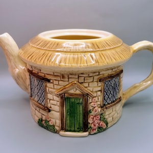 May include: A ceramic teapot shaped like a cottage, featuring a thatched roof and brickwork walls. The teapot has a green door, diamond-patterned windows, and a curved handle. Pink flowers adorn the base, and the spout is shaped like a branch.