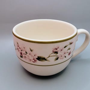 May include: Cream-colored teacup with a floral design. The cup features a delicate pattern of pink flowers and green leaves, with a green trim around the rim and handle. The cup is designed for hot beverages.