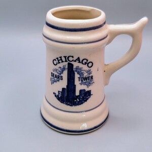 May include: A small, off-white ceramic mug with a handle, featuring a blue illustration of the Chicago skyline, including the Sears Tower. The word "CHICAGO" is arched above the image. Blue stripes accent the top and bottom.