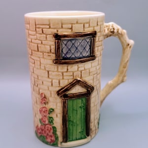 May include: A ceramic mug shaped like a small house with a green door, brown window, and brick exterior. The mug has a tree branch-shaped handle and pink flowers on the side.
