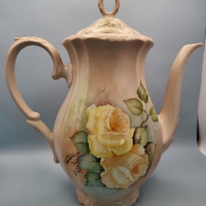May include: A vintage porcelain teapot with a delicate floral design. The teapot is decorated with yellow roses and green leaves on a light pink background. The teapot has a lid with a gold knob.
