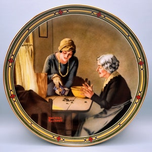 Puede incluir: Un plato de cer&aacute;mica de estilo vintage con una pintura de dos mujeres en un ambiente de cocina. La mujer mayor lleva un vestido oscuro y un delantal blanco, mientras que la mujer m&aacute;s joven lleva un vestido azul y un sombrero blanco. La mujer m&aacute;s joven est&aacute; vertiendo algo de una caja en un taz&oacute;n que sostiene la mujer mayor. El plato tiene un borde dorado y un borde verde y amarillo con un dise&ntilde;o floral rojo. La firma del artista, "Norman Rockwell", es visible en la esquina inferior izquierda del plato.