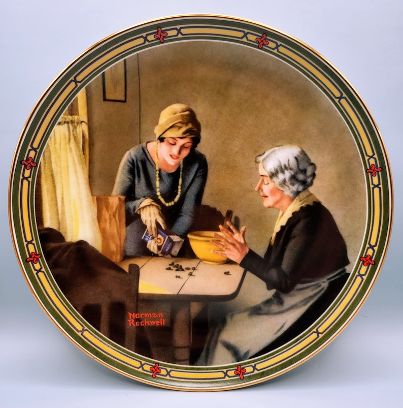 Puede incluir: Un plato de cer&aacute;mica de estilo vintage con una pintura de dos mujeres en un ambiente de cocina. La mujer mayor lleva un vestido oscuro y un delantal blanco, mientras que la mujer m&aacute;s joven lleva un vestido azul y un sombrero blanco. La mujer m&aacute;s joven est&aacute; vertiendo algo de una caja en un taz&oacute;n que sostiene la mujer mayor. El plato tiene un borde dorado y un borde verde y amarillo con un dise&ntilde;o floral rojo. La firma del artista, "Norman Rockwell", es visible en la esquina inferior izquierda del plato.