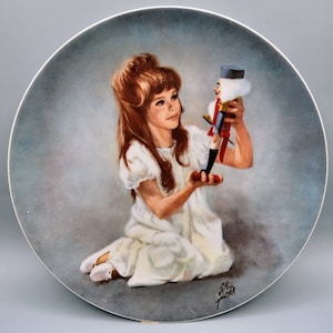 May include: A porcelain plate featuring a painting of a young girl in a white dress holding a wooden nutcracker toy. The girl has long brown hair and is looking at the toy. The background is a light blue colour. The plate is signed "Shell Asher".
