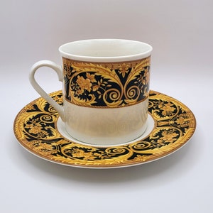 May include: A white porcelain coffee cup and saucer set with a black and gold floral pattern. The cup has a handle and the saucer has a scalloped edge.