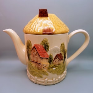 May include: A ceramic teapot with a thatched roof lid and a painted scene of a countryside with houses and trees. The teapot is cream colored with brown, green, and red accents.