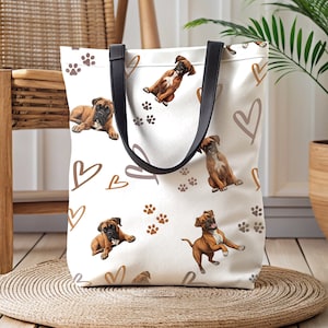 May include: A white canvas tote bag with black handles features a repeating pattern of brown boxer dogs, paw prints, and hearts. The bag is sitting on a woven rug.