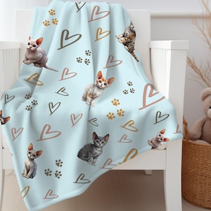 May include: A soft blue fleece blanket with a repeating pattern of brown paw prints, pink hearts, and white Devon Rex cats.
