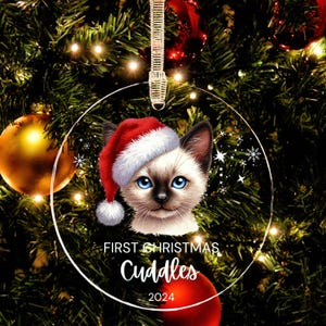 May include: A clear, round Christmas ornament featuring a Siamese cat wearing a Santa hat. The ornament has the text "First Christmas Cuddles 2024" in white script. The background shows a decorated Christmas tree with lights and ornaments.