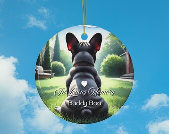 May include: A round ceramic ornament featuring a black French Bulldog sitting with its back to the viewer. The ornament has a white heart on the dog's back and the text "In Loving Memory Buddy Boo" printed on the bottom. The background is a blurred image of a green lawn and a house.