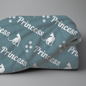 May include: A folded teal blanket with a repeating pattern of white cat silhouettes, paw prints, and the word "Princess" in a cursive font. The blanket is soft and plush, perfect for a cat lover.