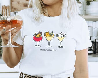 May include: White t-shirt featuring three cartoon cats relaxing in cocktail glasses. The glasses are filled with red, yellow, and white drinks. The text "Happy Cattail Hour" is printed below the glasses. The shirt is worn by a person.