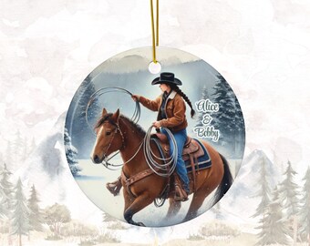 May include: A round ceramic ornament featuring a woman in a cowboy hat riding a brown horse through a snowy winter scene. The woman is holding a lasso and the ornament has the text "Alice & Bobby" on it.