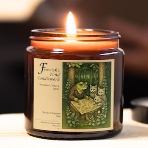 May include: A lit soy wax candle in a glass jar. The label features a frog in a suit, two kittens, and a map. The candle is from the "Woodland Library Series" and is labeled "The Secret Treasure Map".