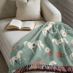 May include: A soft, green throw blanket with a repeating pattern of white horses and floral designs. The blanket has a fringed edge with multiple colors and is draped over a white armchair. An open book and a pillow are also visible.