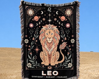 Può includere: Una coperta tessuta nera con un'illustrazione centrale di un leone circondato da motivi celesti tra cui soli, lune, pianeti e stelle. Il testo LEO, CONFIDENT, RADIANT, FEARLESS è stampato in basso con bordi sfrangiati.