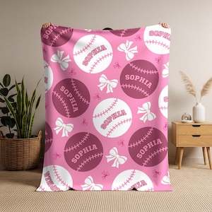 May include: Pink baseball-themed blanket with a repeating pattern of baseballs, bows, and the name "Sophia." The baseballs alternate between white and a darker pink, with white stitching details. The bows are white.