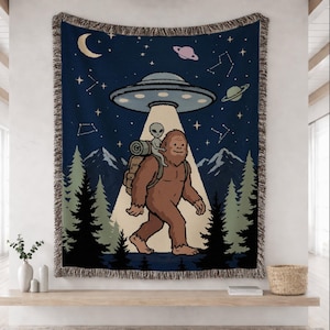 May include: A woven throw blanket featuring a dark blue night sky with a crescent moon, planets, and constellations. A brown Bigfoot carries a small gray alien in a backpack under a light beam from a flying saucer above a forest and mountains.