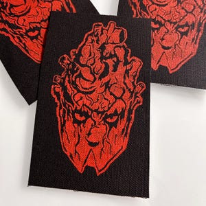 May include: A rectangular black fabric patch featuring a red embroidered graphic of a stylized anatomical heart with a face. The design displays intricate, vein-like textures and sharp, angular features, creating a bold contrast against the black background.
