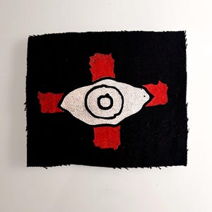 May include: Black square patch with a silver eye design, a red cross, and a black border. The eye has a black outline and a black pupil. The red cross is painted on the black background.