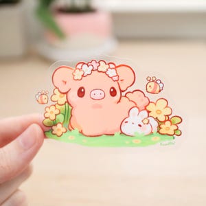 May include: A clear sticker featuring a pink pig with a flower crown, a white bunny, and yellow flowers. There are also two small yellow bees on the sticker.