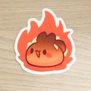 May include: A cartoon sticker of a brown and orange blob with a happy face, surrounded by orange flames. The sticker has a white border and the artist's signature in the bottom right corner.