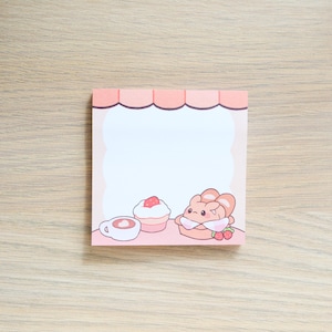 May include: Pink notepad with a white background and a cute cartoon illustration of a pastry, a cupcake, and a cup of coffee. The notepad has a pink awning at the top.