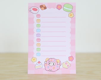 May include: Pink notepad with a checkered pattern and a white cloud border. The notepad has a list of lines for writing and is decorated with images of macarons, a cup of coffee, a piece of cake, and a sheep.