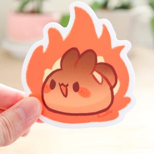 May include: A sticker of a brown cartoon character with a happy expression, set against a background of orange flames. The character has a round body and small arms and legs. The sticker is white with a white border.