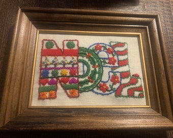 May include: Framed embroidery art spelling out the word "JOY". The letters are stitched with various colors and patterns, including red, green, and white. The frame is made of wood.