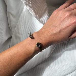 Può includere: Un bracciale a fascia in metallo color bronzo dal design aperto, caratterizzato da un piccolo fiore scolpito su un'estremità e una piccola ape scolpita sull'altra. Il bracciale presenta una finitura liscia e lucida attorno al polso.