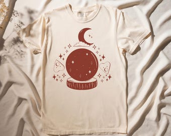 May include: A white t-shirt with a red illustration of a crystal ball, crescent moon, and stars. The crystal ball is on a wooden stand.