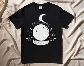 May include: Black t-shirt with a white graphic of a crystal ball, crescent moon, and stars. The graphic is in a vintage style.