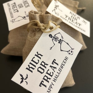Puede incluir: Tres peque&ntilde;as bolsas de arpillera con etiquetas blancas atadas a ellas. Las etiquetas tienen un gr&aacute;fico de esqueleto negro y el texto "Kick or Treat Happy Halloween!"