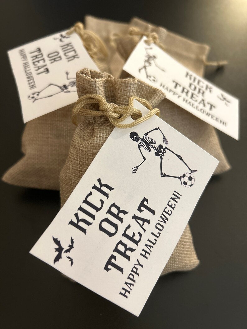 Puede incluir: Tres peque&ntilde;as bolsas de arpillera con etiquetas blancas atadas a ellas. Las etiquetas tienen un gr&aacute;fico de esqueleto negro y el texto "Kick or Treat Happy Halloween!"