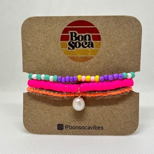 May include: A set of three colorful beaded bracelets with a pearl charm. The bracelets are layered in a rainbow pattern of pink, purple, yellow, white, and turquoise. The charm is attached to the bottom bracelet with a gold chain. The bracelets are displayed on a brown card with the text "Bon Soca" and "@bonsocavibes".