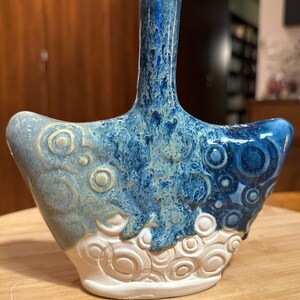 May include: A blue and white ceramic vase with a unique, abstract design. The vase has a wide base and a narrow neck, and it is decorated with swirling patterns and circles.