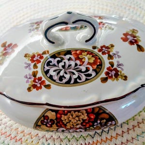 Puede incluir: Fuente ovalada blanca con tapa, adornada con motivos florales en rojo, naranja y morado. Presenta una pieza central floral negra y dorada y asas curvas. Un artículo de menaje decorativo.