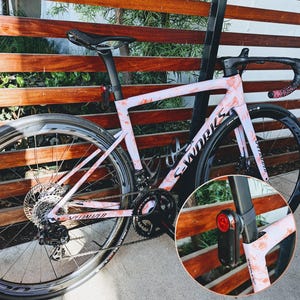Puede incluir: Una bicicleta de carretera Specialized S-Works con un estampado floral rosa y rojo. La bicicleta tiene neum&aacute;ticos negros, un asiento negro y una luz trasera. El cuadro tiene el logotipo "S-Works". La bicicleta est&aacute; apoyada en una valla de madera.