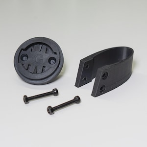 Puede incluir: Un kit de montaje para ordenador de bicicleta negro. El kit incluye una base redonda con la letra "G" en el centro, un soporte curvo y dos tornillos negros. Las piezas son de material negro mate.