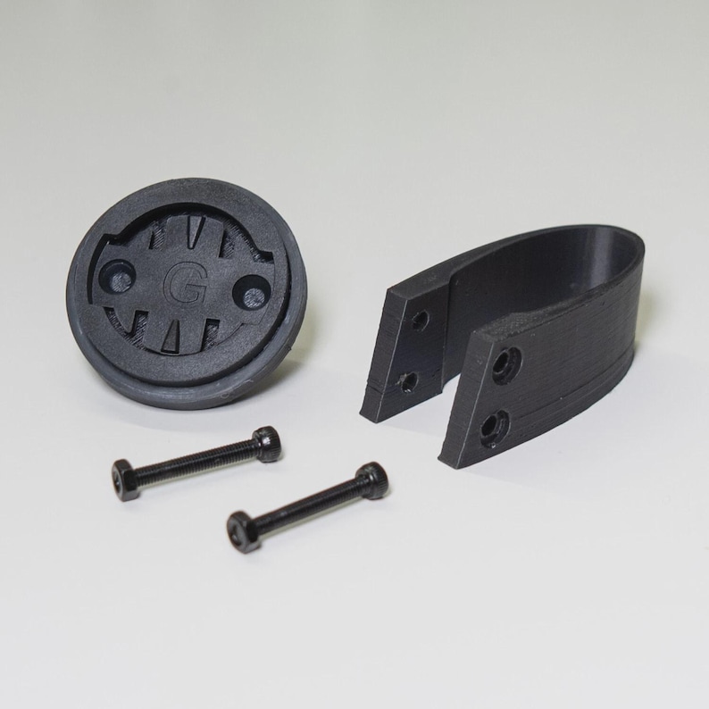Puede incluir: Un kit de montaje para ordenador de bicicleta negro. El kit incluye una base redonda con la letra "G" en el centro, un soporte curvo y dos tornillos negros. Las piezas son de material negro mate.