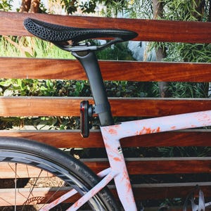 Puede incluir: Bicicleta de carretera con sill&iacute;n y tija negros, y cuadro claro con detalles rojos. Una luz roja y negra est&aacute; sujeta a la tija. Se ve la rueda de la bicicleta.