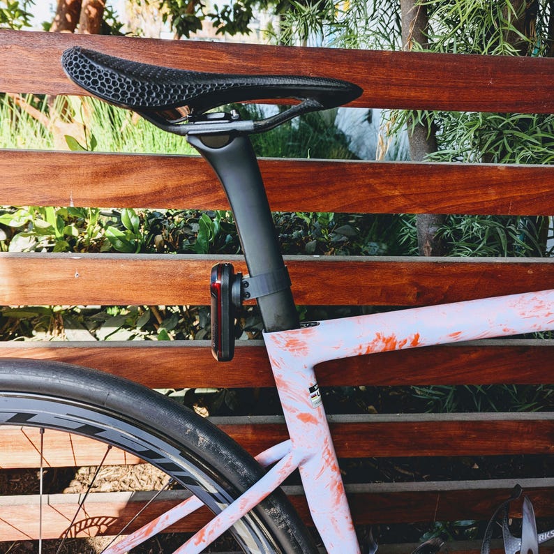 Puede incluir: Bicicleta de carretera con sill&iacute;n y tija negros, y cuadro claro con detalles rojos. Una luz roja y negra est&aacute; sujeta a la tija. Se ve la rueda de la bicicleta.