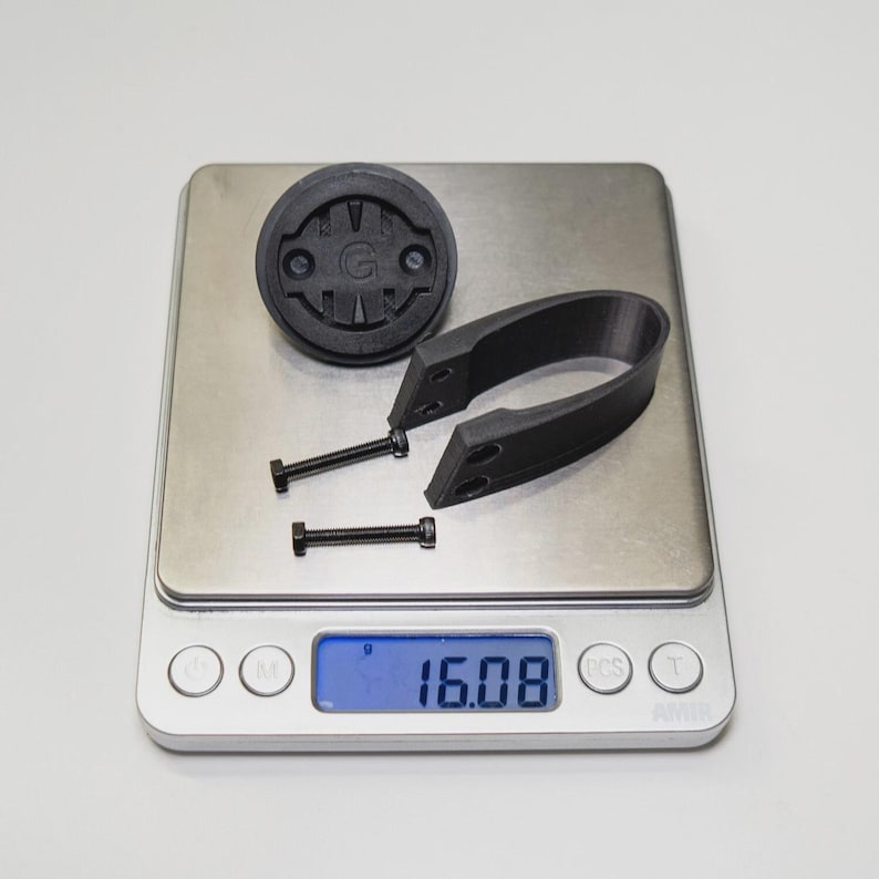 Puede incluir: Un soporte y un soporte negros para ordenador de bicicleta con dos tornillos, y una pieza redonda negra con la letra "G" se muestran en una balanza digital. La balanza indica 16,08, lo que probablemente indica el peso en gramos.