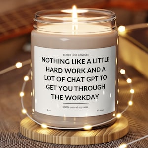 May include: A brown soy wax candle in a clear glass jar with a white label that reads "NOTHING LIKE A LITTLE HARD WORK AND A LOT OF CHAT GPT TO GET YOU THROUGH THE WORKDAY". The candle is on a wooden coaster and surrounded by string lights.