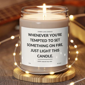 Könnte beinhalten: Eine braune Sojawachskerze mit einem weißen Etikett, auf dem steht "Whenever you're tempted to set something on fire, just light this candle." Die Kerze steht auf einem Holzuntersetzer und ist von weißen Lichterketten umgeben.