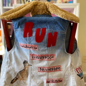 May include: A light blue denim vest with the word "RUN" in red felt letters on the back. The vest has patches with the names of states, including Alabama, Mississippi, Tennessee, and Arizona. There is also a bird patch on the left side of the vest.