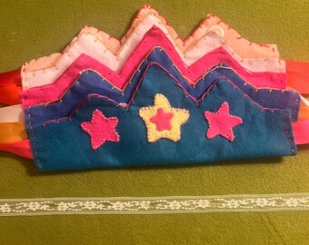 May include: A handmade felt crown in shades of blue, pink, and white, with star embellishments. The crown has a zigzag design and is accented with gold stitching and colorful ribbons. A white lace ribbon is also visible.