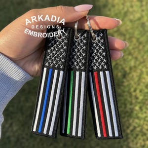 May include: Embroidered keyrings with an American flag design. Each keyring has a black background with white stars and stripes, and a coloured stripe in blue, green, or red. The text "ARKADIA DESIGNS EMBROIDERY" is visible.